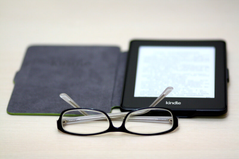 A kindle placed flat on a surface with its cover open turned on with text on the screen. a pair of glasses lay in front.